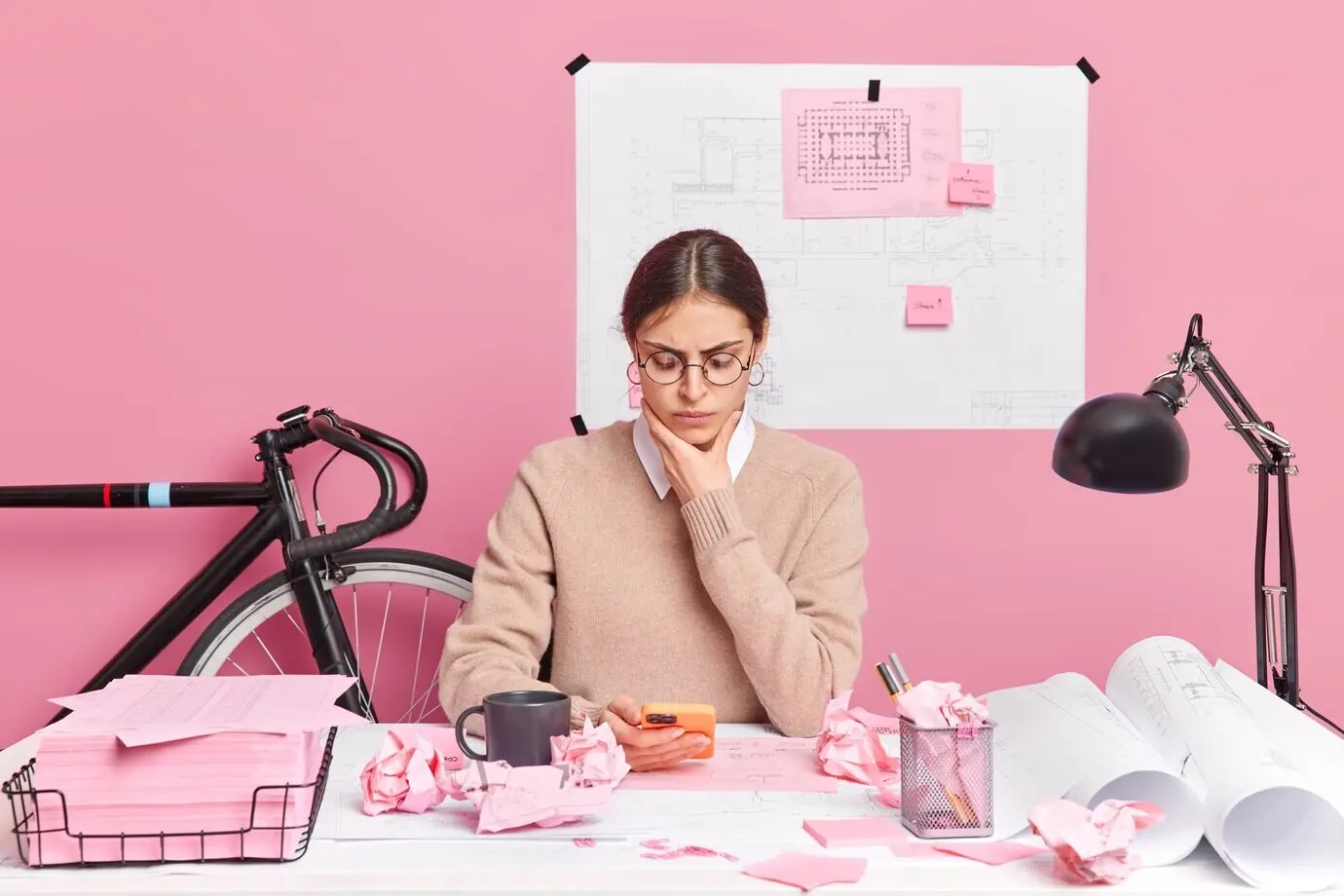 A serious young woman wearing glasses creates sketches and blueprints in an office using a smartphone, posing at a desk against a pink wall. A professional graphic designer develops a new strategy.