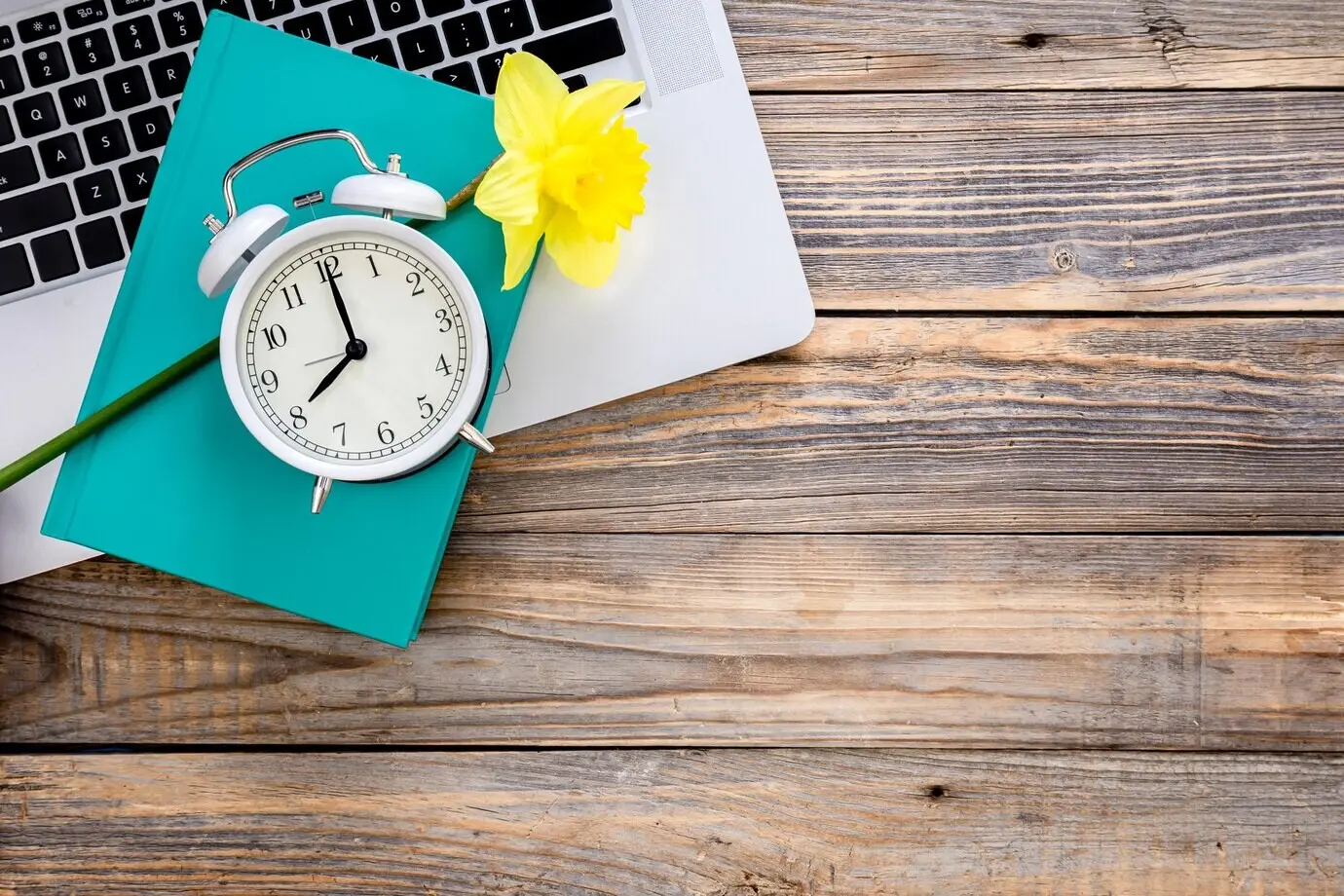 Top view of a laptop, a book, a flower, and a white alarm clock on a wooden background.