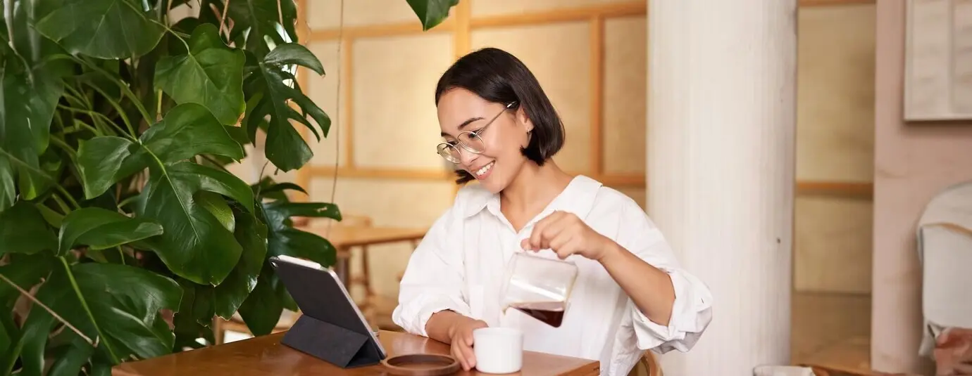 A smiling young woman, a freelance and remote worker, pouring coffee into a cup while sitting in a cafe and looking.